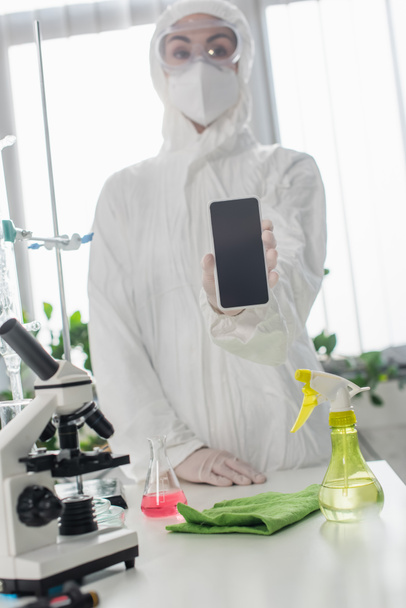 研究室の顕微鏡の近くにスマートフォンがあるのを示す個人用保護具のぼやけた科学者 - 写真・画像