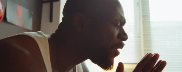 Κοντινό πλάνο κλίση προς τα κάτω πυροβολισμό του Αφροαμερικανού άνδρα κάθεται με τα μάτια κλειστά, κρατώντας τα χέρια σφιγμένα και λέγοντας προσευχή στο σπίτι - Πλάνα, βίντεο