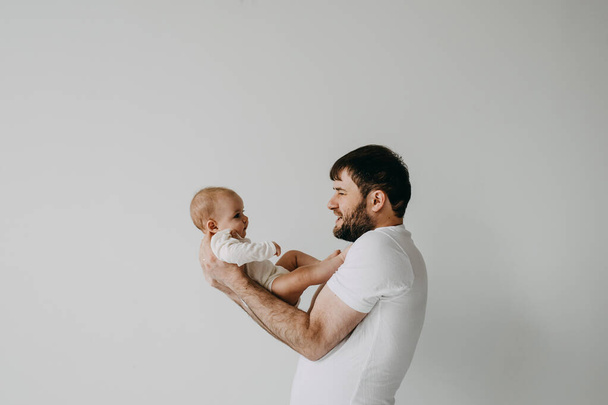 Πατέρας κρατώντας το μωρό στην αγκαλιά, χαμογελώντας, σε λευκό φόντο τοίχο. - Φωτογραφία, εικόνα
