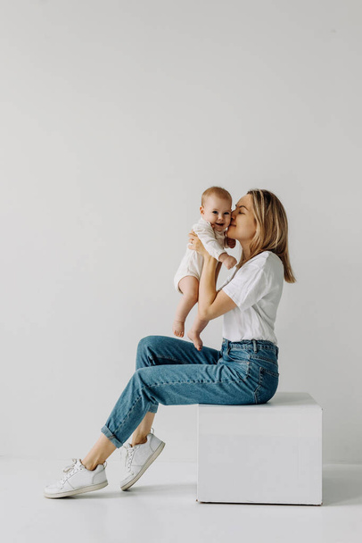 Mother sitting and holding baby in arms, smiling and playing, on white wall background. - Foto, Imagen