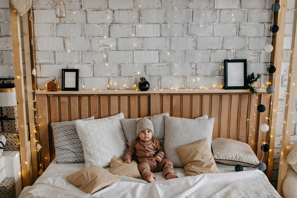 Baby sitting on bed between pillows at home, barefoot, wearing a beanie with ears. - Foto, Imagem