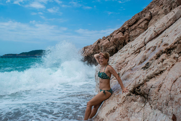 Woman stands near rocks in surf on white spray large sea waves background. Kamenovo pebble beach, located near Rafailovici village. Sunny summer day. Budva Riviera. Montenegro. - Foto, imagen