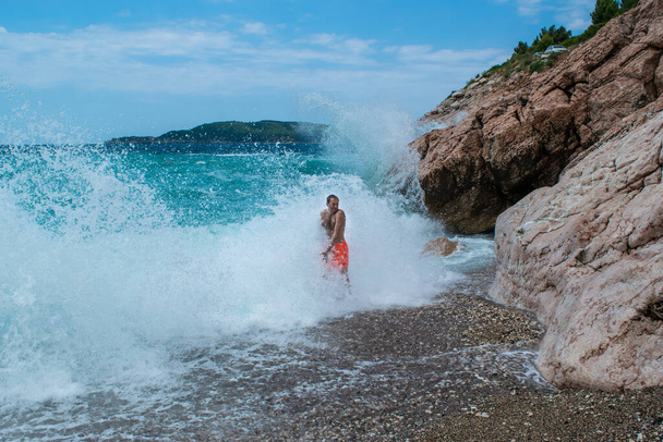 Hombre se encuentra en spray blanco de grandes olas marinas cerca de rocas en la playa de guijarros de Kamenovo, situado cerca de Rafailovici pueblo. Día soleado de verano. Budva Riviera. Montenegro. - Foto, imagen
