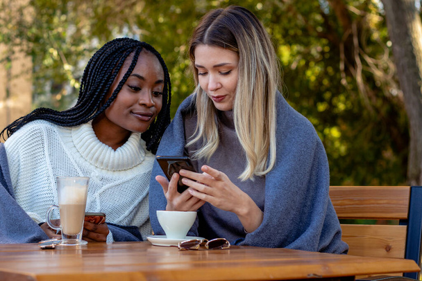 Two young girlfriends enjoy coffee together on a terrace of the coffee shop, sitting at a table, chatting on social networks, and laughing. Diversity and multi-ethnic concept. - Φωτογραφία, εικόνα