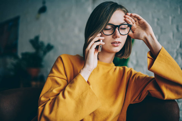 Tired female in casual clothing and optical spectacles feeling headache while received bad news during cellphone conversation, unhappy Caucaisan woman using wireless connection for phone calling - Foto, afbeelding