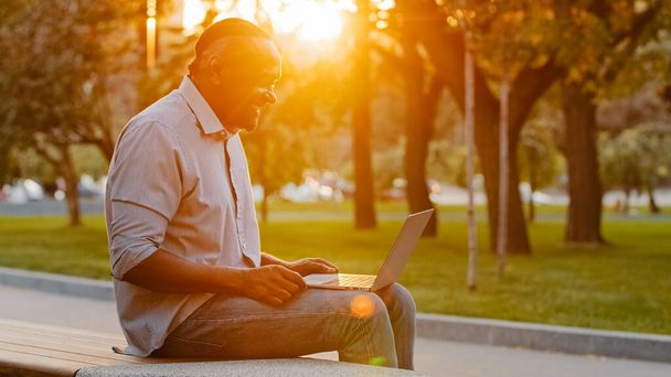 Yaşlı Afro-Amerikan adam dışarıda oturuyor modern cihazda kablosuz internete bakıyor, dizüstü bilgisayarda mesaj yazıyor, yetişkin serbest yazar müşteriye danışmanlık yapıyor, teknoloji konsepti - Fotoğraf, Görsel