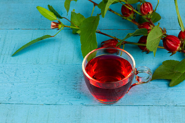 Roselle tea or Hibiscus sabdariffa juice in transparent glass and fresh roselle on blue wooden background. Drink herbal water for health care. - Fotoğraf, Görsel