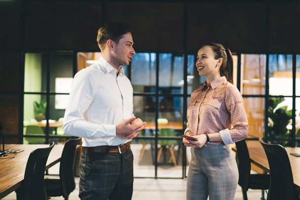 Positive man and woman in formal outfits standing next to each other and discussing business project while working in modern office - Foto, afbeelding