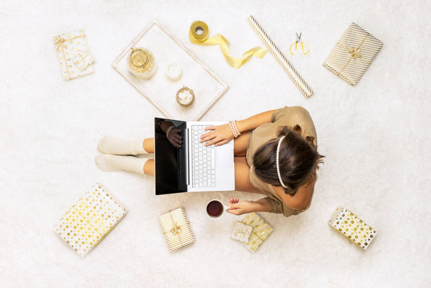 Christmas. Woman in sweater using laptop for searching gift ideas sitting on the white carpet among the many wrapped boxes in white gold wrapping paper. Top view. Concept - Foto, Imagem