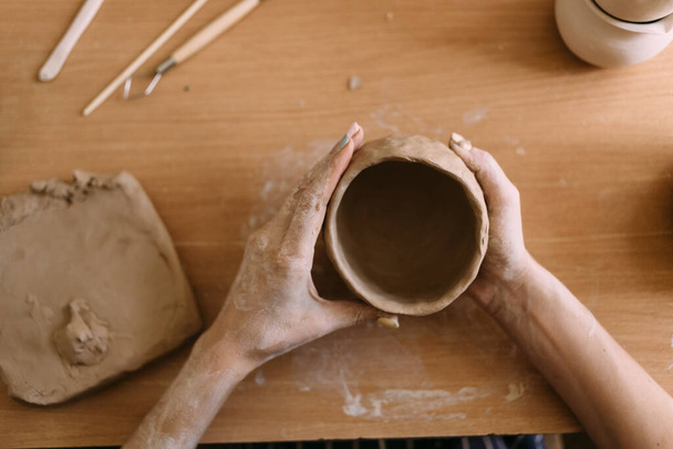 陶芸のワークショップ粘土芸術の概念。焼き立てのない土鍋を持った若い陶芸家の手によるクローズアップ。職人の手は粘土コップと装飾道具を持っています。テキストの場所 - 写真・画像
