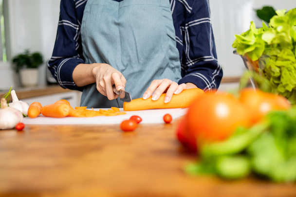 Close up hand of young lady woman preparing vegetables for food salad in kitchen by slicing carrot and chop on chopping board - Fotó, kép