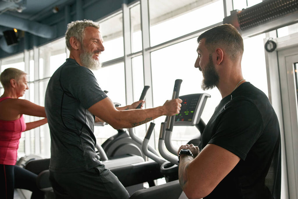 Vrolijke volwassen man gelukkig chatten met trainer tijdens het sporten - Foto, afbeelding