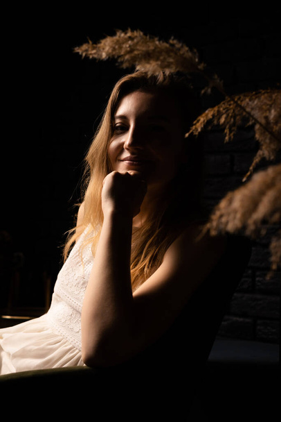 A young woman in a white dress sits thoughtfully in an armchair in a dark room. Solitude, introspection concept. - Foto, Imagem