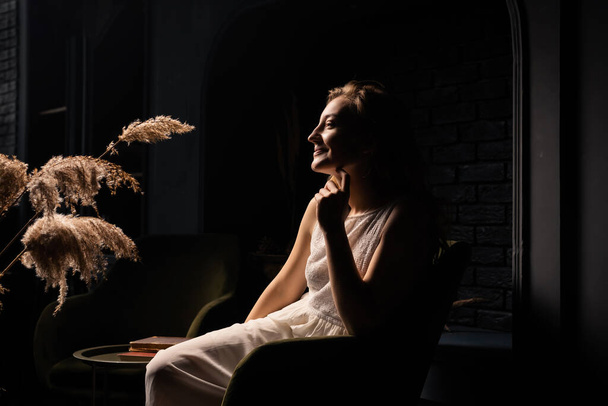 Een jonge vrouw in een witte jurk zit bedachtzaam in een fauteuil in een donkere kamer. Eenzaamheid, introspectie concept. - Foto, afbeelding