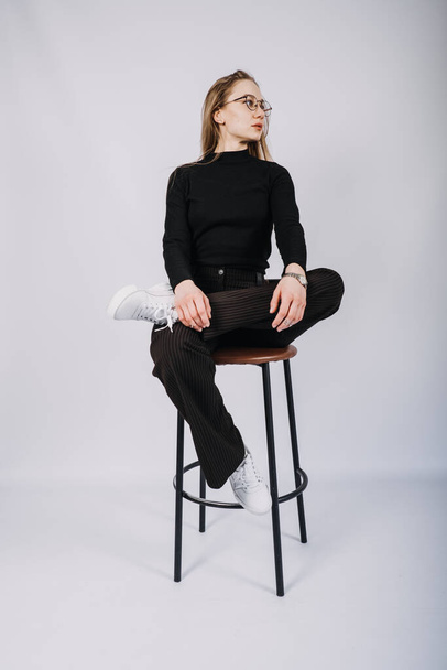 Young woman, fashion designer, stylist full-length casual style portrait. Fashionable woman in black turtleneck and bar stool. Studio shot of stylish blonde girl with long hair in casual clothes. - Fotó, kép