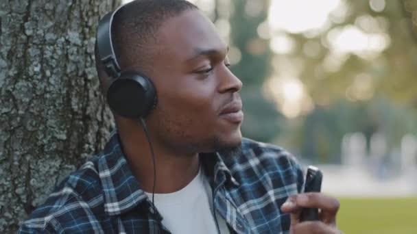 Male portrait african american young guy funny man in headphones listening to music using phone online radio pretending play drums sings in smartphone like microphone enjoying song audio sound in park - Footage, Video