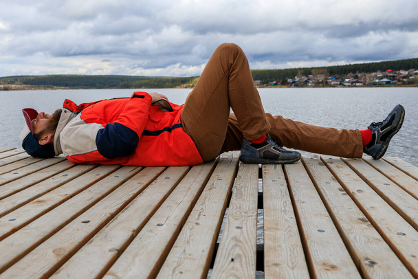 a bearded man in a red jacket and a cap is resting on a wooden pier. rest outside the city, calm down by the water - Photo, Image
