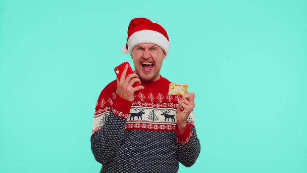 Ο άνθρωπος στα Χριστούγεννα κόκκινο πουλόβερ χρησιμοποιούν κινητό τηλέφωνο και πλαστικό πιστωτική κάρτα τράπεζα, νίκη, calebrate, wow - Φωτογραφία, εικόνα