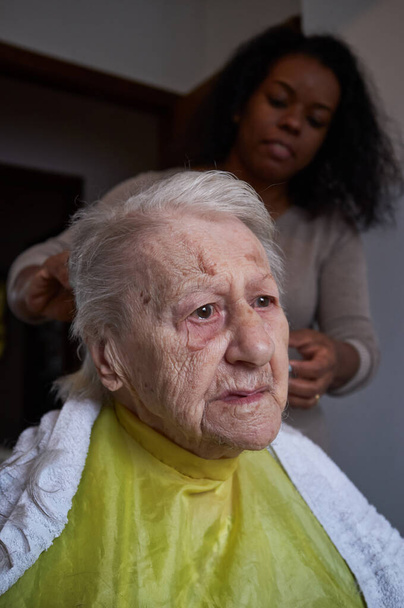 Verzorgende Afro-Amerikaanse vrouw verzorger, knippen van haar bejaarde vrouw haar op hom - Foto, afbeelding