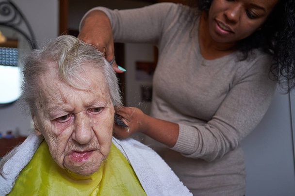 Fürsorgliche afroamerikanische Pflegerin, die ihrer älteren Frau die Haare schneidet - Foto, Bild