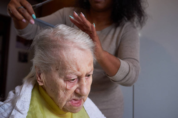 Huolehtiva Afrikkalainen Amerikkalainen nainen hoitaja, leikkaus hänen iäkäs nainen hiukset hom - Valokuva, kuva
