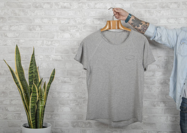 Tetovaná mužská ruka drží ramínko s šedou špičkovou kvalitou jemného bavlněného trička. Vložit reklamu pro kopírování Mockup - Fotografie, Obrázek