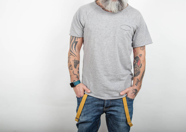 Bearded tattooed man poses in jeans with suspenders and gray short-sleeved cotton t-shirt, on white background. - Photo, Image