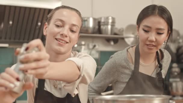 Primo piano medio di giovani donne asiatiche e caucasiche che indossano grembiuli, in piedi vicino ai fornelli in cucina professionale, condimento di cibo invisibile, parlare e sorridere - Filmati, video
