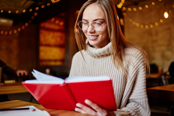 Crop satisfied female student in casual sweater sitting at table in cafe and reading notes in notebook while doing homework - Foto, imagen