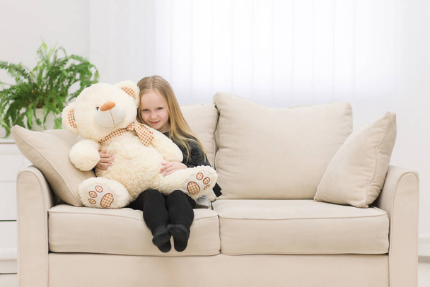 Little blonde girl sitting on the sofa and hugging her teddy bear photo. - Foto, Bild
