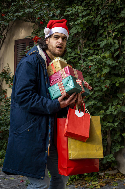 Время покупок на Рождество. Молодой человек в шляпе Санта-Клауса имеет удивленное выражение лица и приносит свои недавно приобретенные подарки. - Фото, изображение