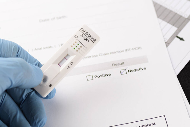Detailní záběr negativního výsledku rychlým antigenovým testem na SARS-CoV-2, COVID-19, který je držen v ruce nad dokumentačním papírem. COVID-19, testování, medicína, pandemie a bezpečnostní koncepce - Fotografie, Obrázek