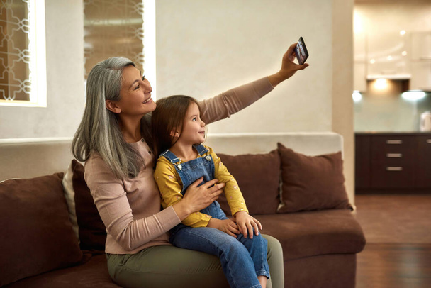 Cheerful girl and grandmother taking selfie at home - Foto, afbeelding