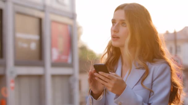Egy lány gyönyörű üzleti nő szőke felhasználó áll a napsugarak napnyugta nézi a telefont a mobil alkalmazás navigáció online térkép keresés rámutatva ujjal maga előtt beszél - Felvétel, videó
