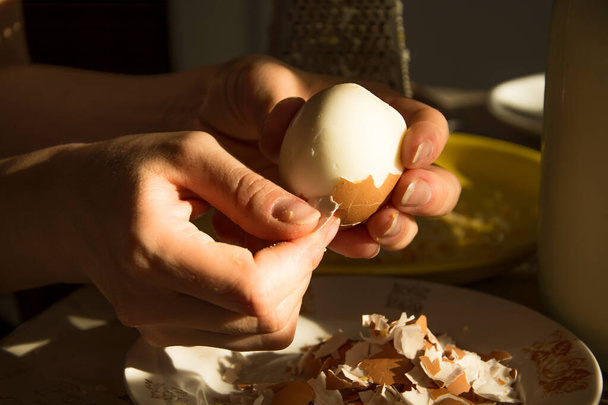 Γυναίκα αφαιρώντας το κέλυφος αυγού από μια βραστή φωτογραφία αρχείου αυγού. Φλούδα αυγού στο τραπέζι της κουζίνας, υλικά για το πιάτο, μαγείρεμα. - Φωτογραφία, εικόνα