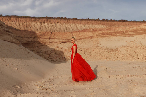 Volledige afbeelding van een blonde jonge vrouw in rood golvende jurk met vliegende stof, kijken naar de camera, poseren op de achtergrond van gouden zand. - Foto, afbeelding