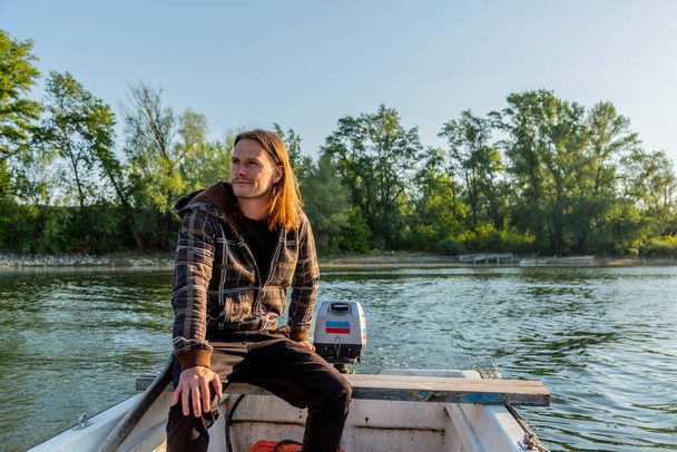 Cool white man with long brown hair is riding white boat with outboard engine in morning. River bank with trees is in background and water is steady. He seems emotional and happy. - Foto, immagini