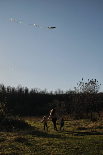 Мама, син і дочка біжать через поле на заході сонця і запускають повітряного змія в синє небо. Приємно проводити час з дітьми на свіжому повітрі. Передній вид. - Фото, зображення