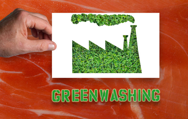 Greenwashing: Een man 's hand houdt een wit vel papier met de omtrek van een fabriek uitgesneden met groen gras binnen.Achtergrond met ham vlees. Niet alles wat 'groen' is, is milieuvriendelijk - Foto, afbeelding