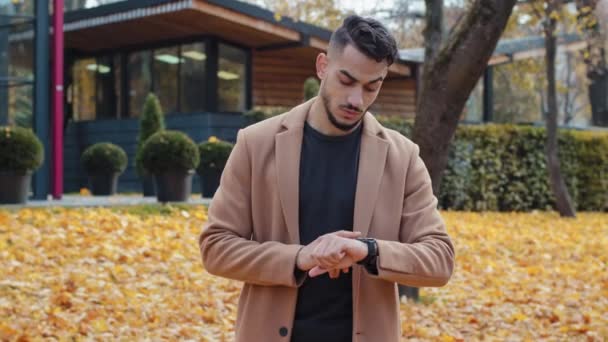 Young impatient hispanic handsome guy in autumn park man look around waiting for meeting discontentedly looking at wrist watch checks time and shakes head angry bearded male nervous standing outdoors - Footage, Video