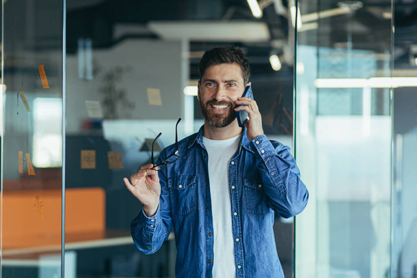 Smiling young adult bearded hipster professional business man making a business call while talking on the phone in the office, enjoying a corporate mobile conversation indoors, close up - Foto, immagini
