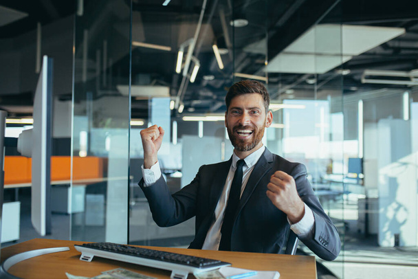 Handsome joyful young businessman sitting at a desk, working in a modern office, throwing money over his head. Happy smiling man throws money in the air - Photo, Image