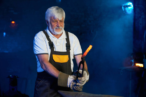 Mature blacksmith working with heated steel on anvil - Fotó, kép