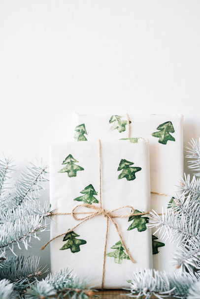 Новогодний подарок упакован в праздничную бумагу с акварелью Рождественские елки на деревянном столе против белой стены с белыми еловыми ветвями - Фото, изображение