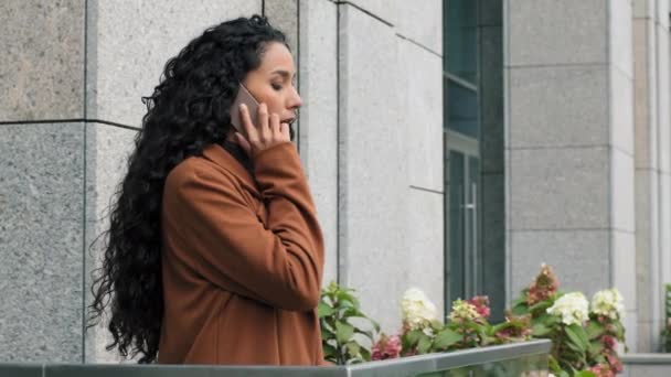 Side view beautiful young confident pensive sad brunette girl with long hair in brown coat business woman going to balcony at home in office talking on phone distant mobile conversation answering call - Footage, Video