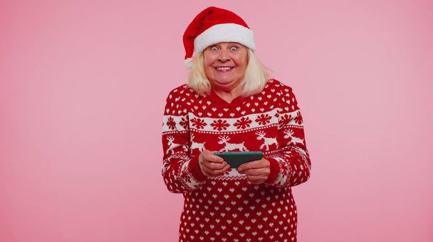 Grand-mère en pull de Noël à la recherche d'affichage smartphone se réjouissant sincèrement gagner de la chance de succès - Photo, image