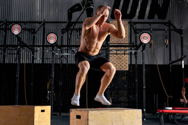 Young caucasian shirtless male is jumping on cross fit wooden box, at gym - Foto, afbeelding