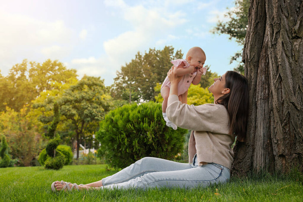 Happy mother with adorable baby sitting on green grass in park - Фото, зображення
