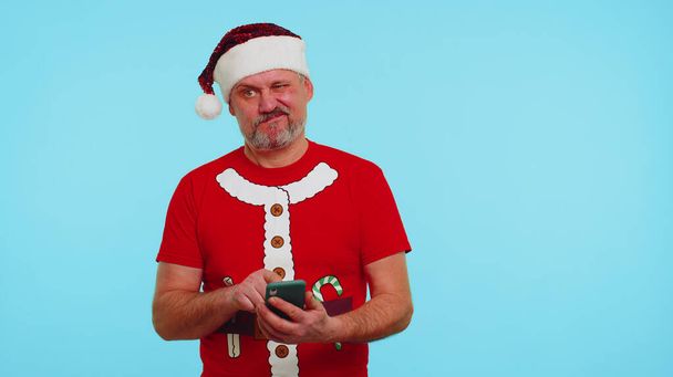 Man in Christmas t-shirt, καπέλο χρησιμοποιώντας κινητό τηλέφωνο πληκτρολογώντας νέα θέση στο διαδίκτυο, online ψώνια, περιήγηση - Φωτογραφία, εικόνα