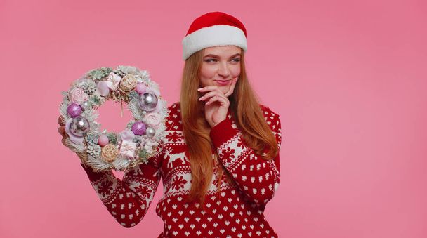 Взрослая улыбающаяся девушка в красном рождественском свитере держит указательный палец на праздничной игрушке - Фото, изображение